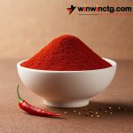 A bowl of vibrant red chili powder, with a fresh chili pepper beside it and scattered peppercorns on a textured brown surface.