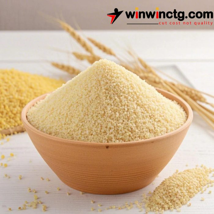 A brown bowl filled with finely ground couscous, with spilled grains nearby and wheat stalks in the background.