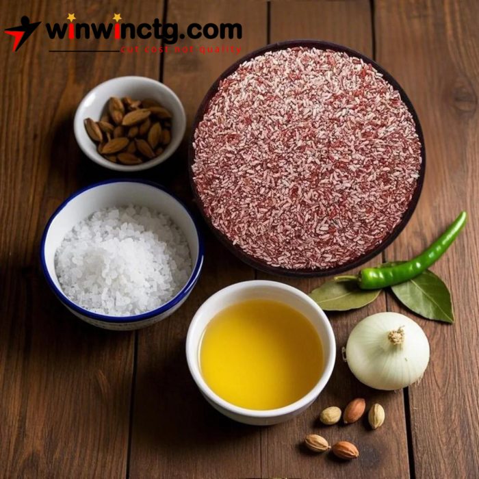 A wooden table displays a bowl of red rice, salt, oil, green chili, onion, and spices, arranged for cooking preparation.