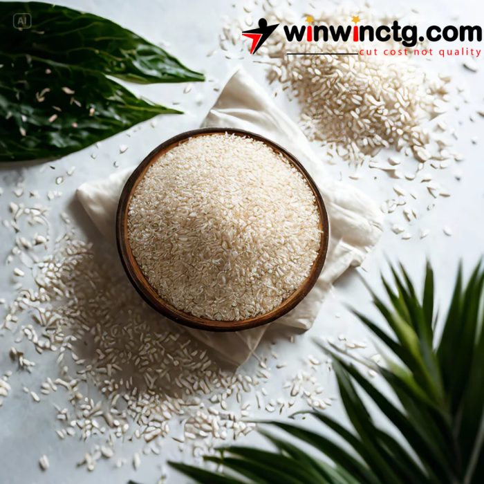 A bowl of uncooked WHITE BINNI RICE on a cloth with rice grains scattered around, surrounded by green leaves. A logo is visible in the top right corner.